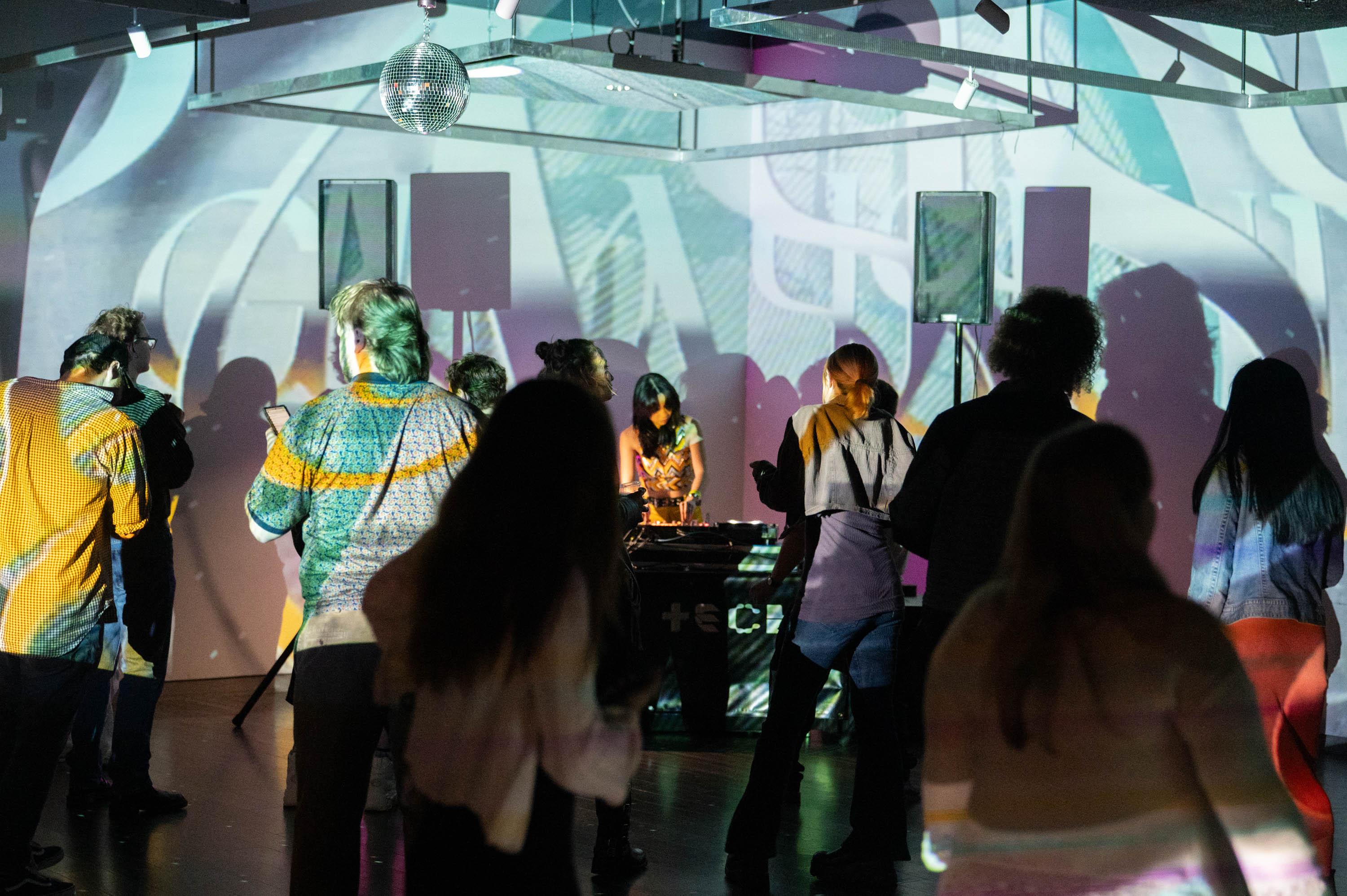 a crowd dances in front of a DJ who has art projected on the wall behind her
