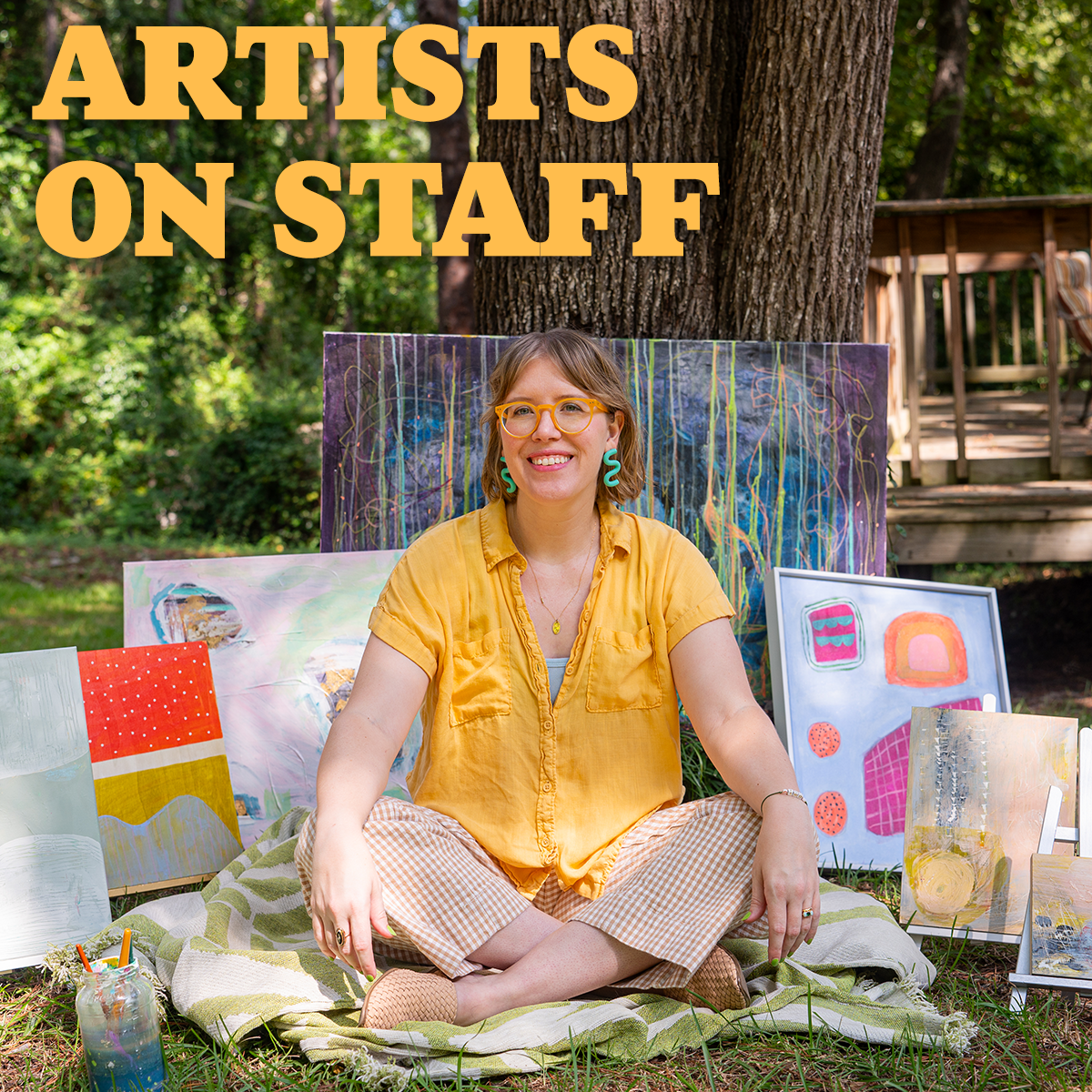 katie sits cross-legged on a blanket in front of a tree, surrounded by her art